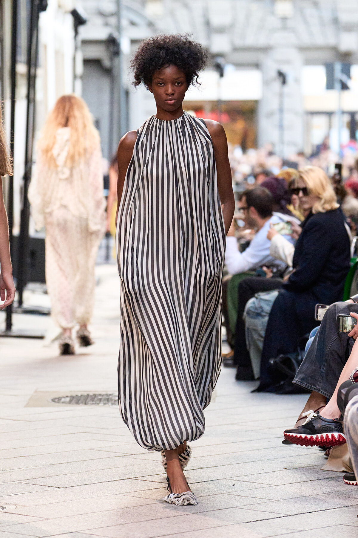 Model walks a street fashion show in a sleeveless black-and-white striped halter maxi dress, audience on both sides.
