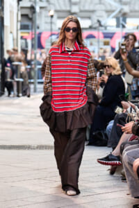 Model walking a city street fashion show in a red and white striped polo top with plaid sleeves and a brown voluminous skirt, wearing sunglasses, surrounded by spectators.