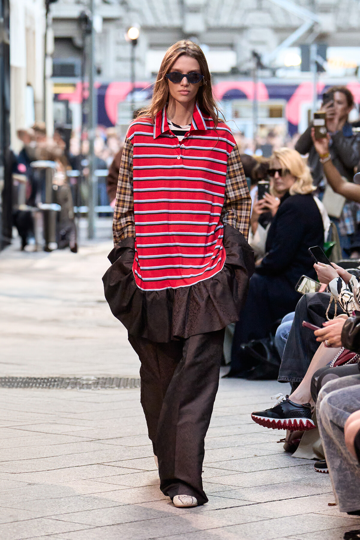 Model walking a city street fashion show in a red and white striped polo top with plaid sleeves and a brown voluminous skirt, wearing sunglasses, surrounded by spectators.
