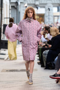 Model walks a pink snakeskin-pattern coat down a street runway, hands in pockets, spectators watching nearby.