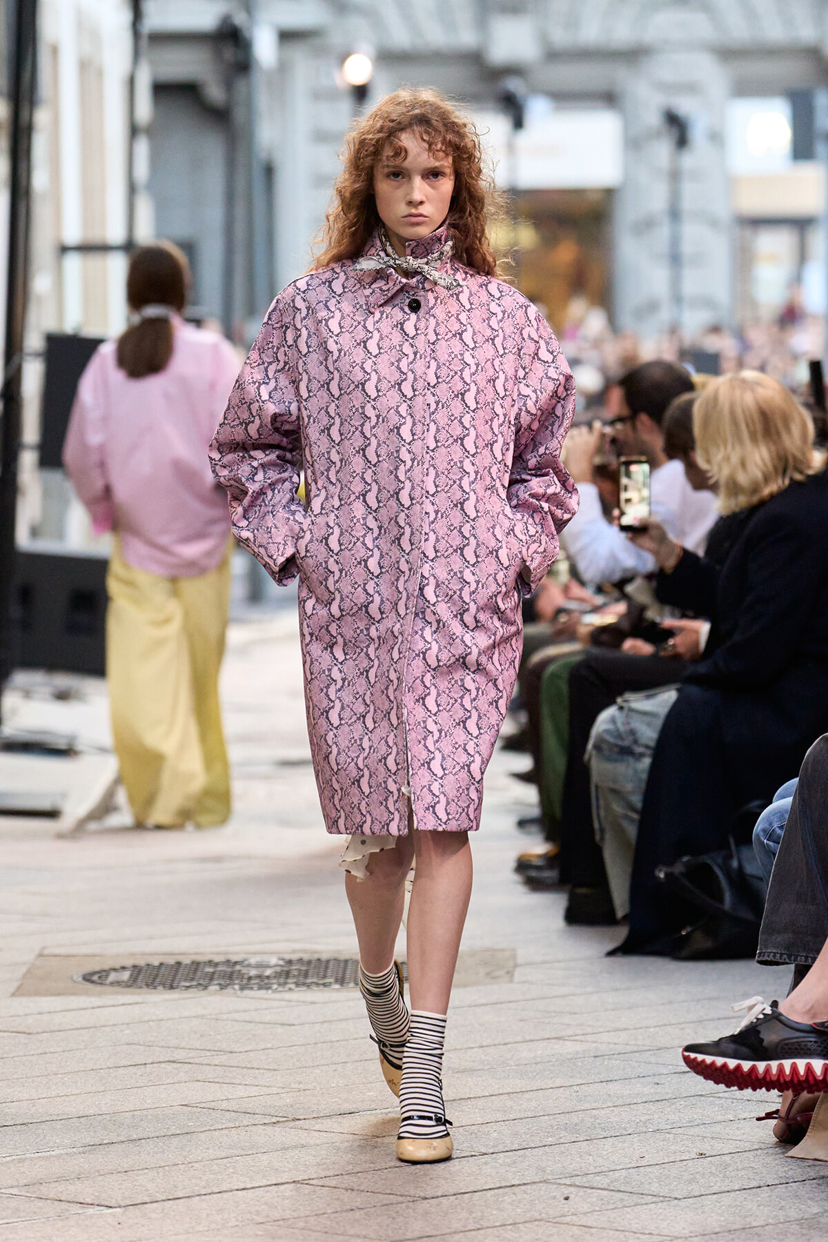 Model walks a pink snakeskin-pattern coat down a street runway, hands in pockets, spectators watching nearby.