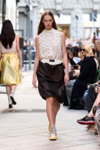 Female model walks a city street runway in a white polka-dot sleeveless blouse and black voluminous skirt, audience watchers in background.