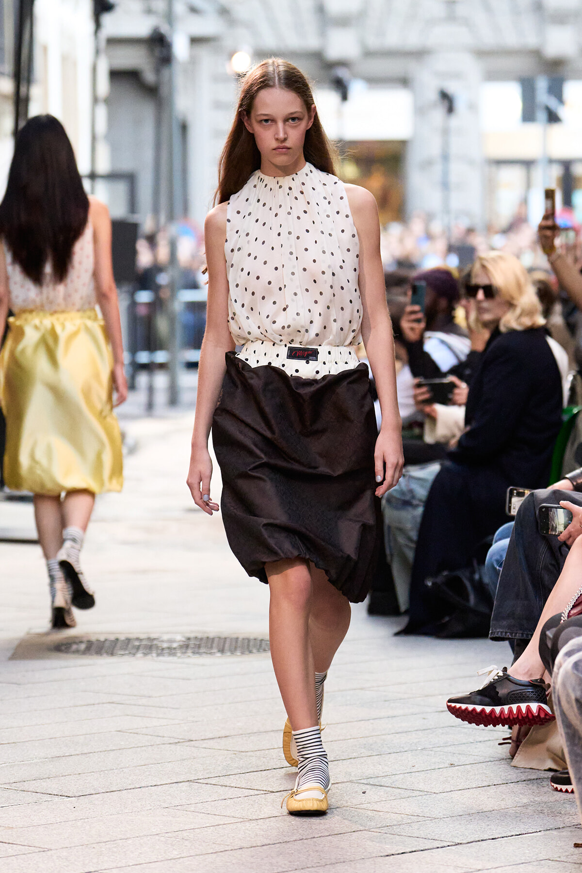 Female model walks a city street runway in a white polka-dot sleeveless blouse and black voluminous skirt, audience watchers in background.