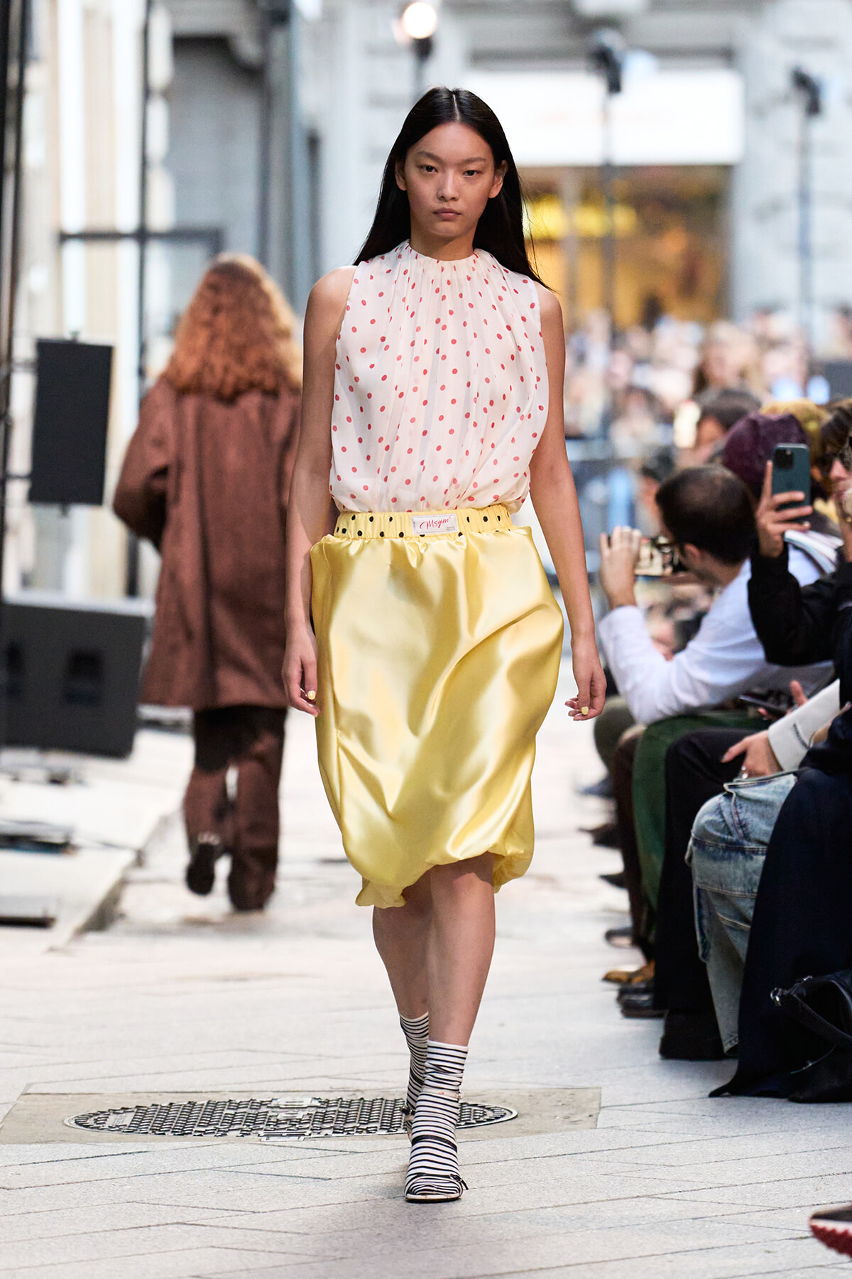 Model walking a fashion show in a white sleeveless blouse with red dots and a shiny yellow bubble skirt; audience watches from sides.