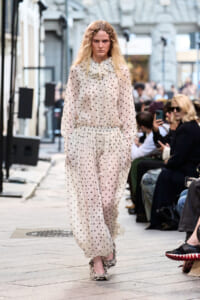 Model walking a street-style fashion show in a sheer cream polka-dot blouse and matching wide-leg pants, audience seated along the runway.