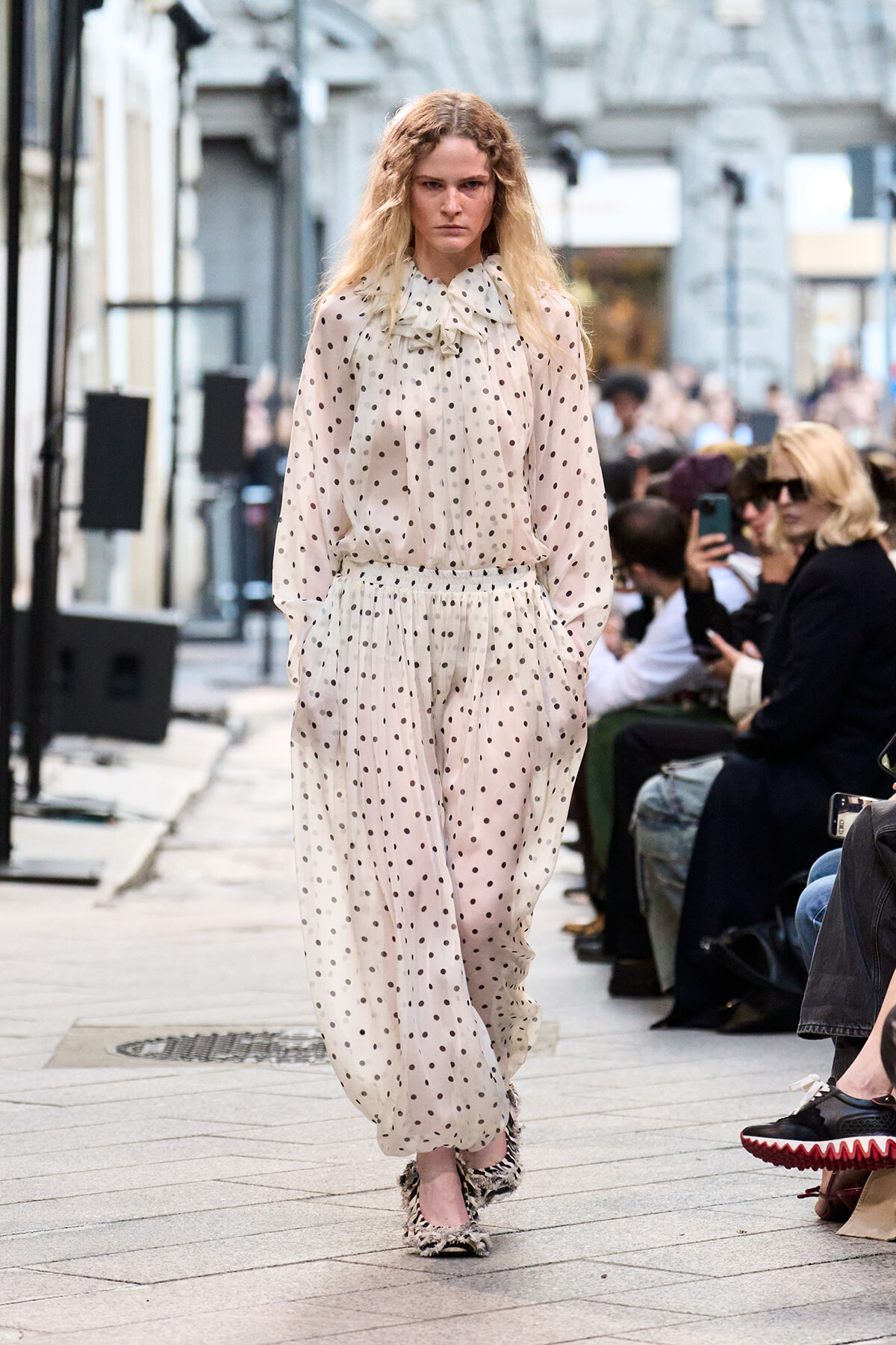 Model walking a street-style fashion show in a sheer cream polka-dot blouse and matching wide-leg pants, audience seated along the runway.