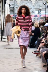 Model walking runway in a burgundy striped polo, light pink knee-length skirt, carrying a tan bag, on a city street with spectators.