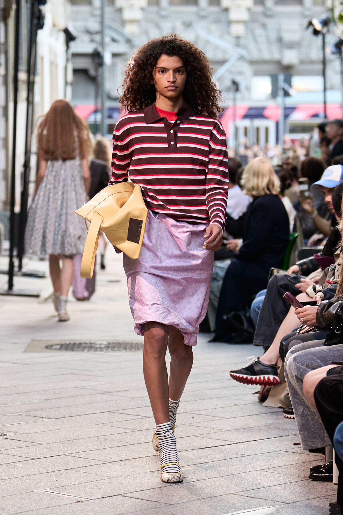 Model walking runway in a burgundy striped polo, light pink knee-length skirt, carrying a tan bag, on a city street with spectators.