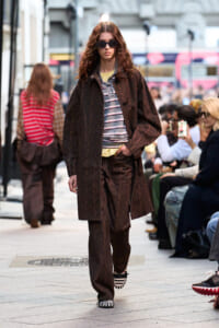 Model walks a city street runway in a dark brown patterned coat and matching trousers, striped top, sunglasses, and striped shoes amid an audience