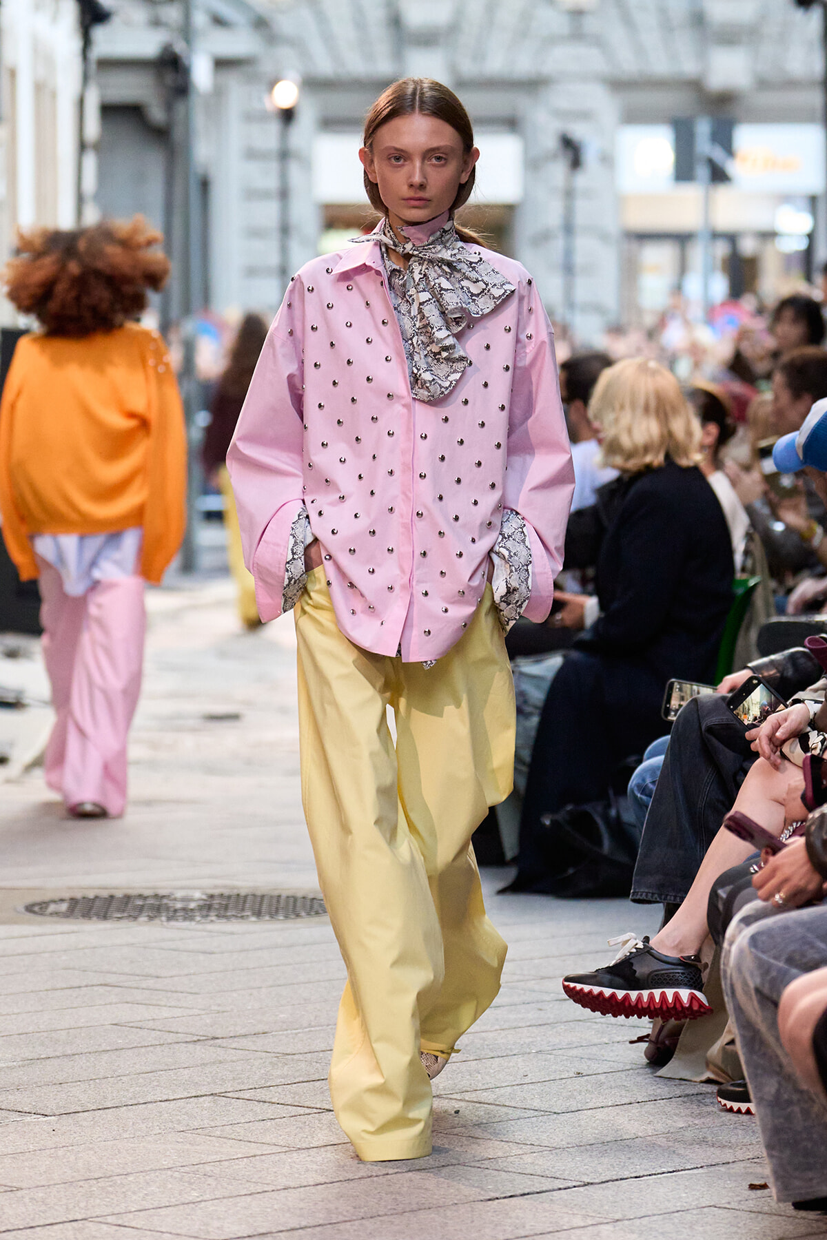 Model walking a street-style runway in a pink studded shirt, snake-print scarf, and loose yellow pants with a crowd watching.