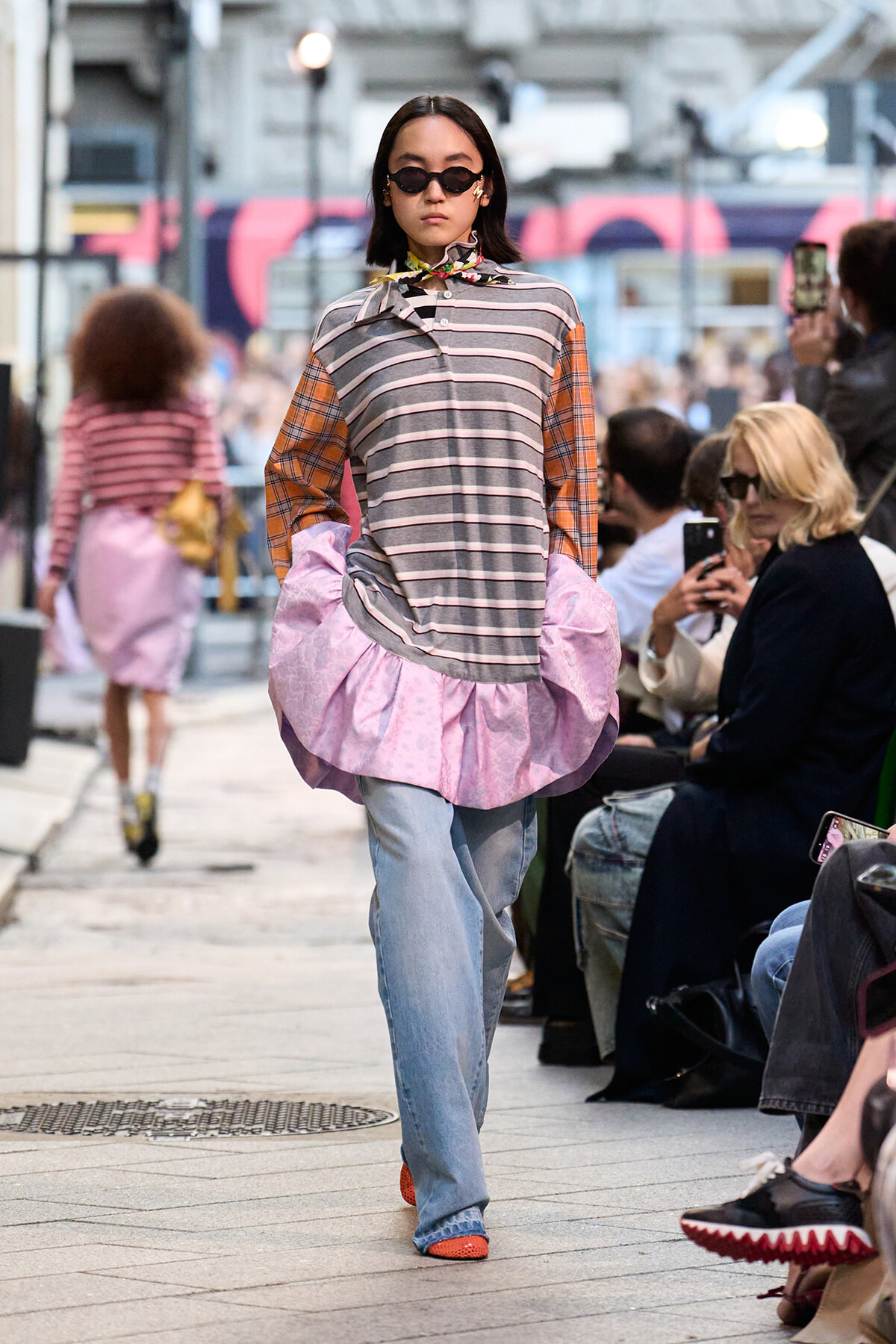 Model walks runway in a striped top with orange sleeves and a pink ruffled hem, wearing jeans and sunglasses in a city street show.
