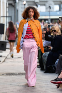 Model with curly hair walks a city street runway wearing an orange studded cardigan over layered striped tops and wide pink pants, with a scarf.