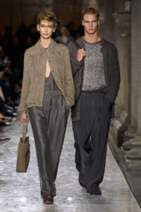 Two models walk a runway; woman in a textured brown cardigan with gray wide-leg pants and a taupe tote.