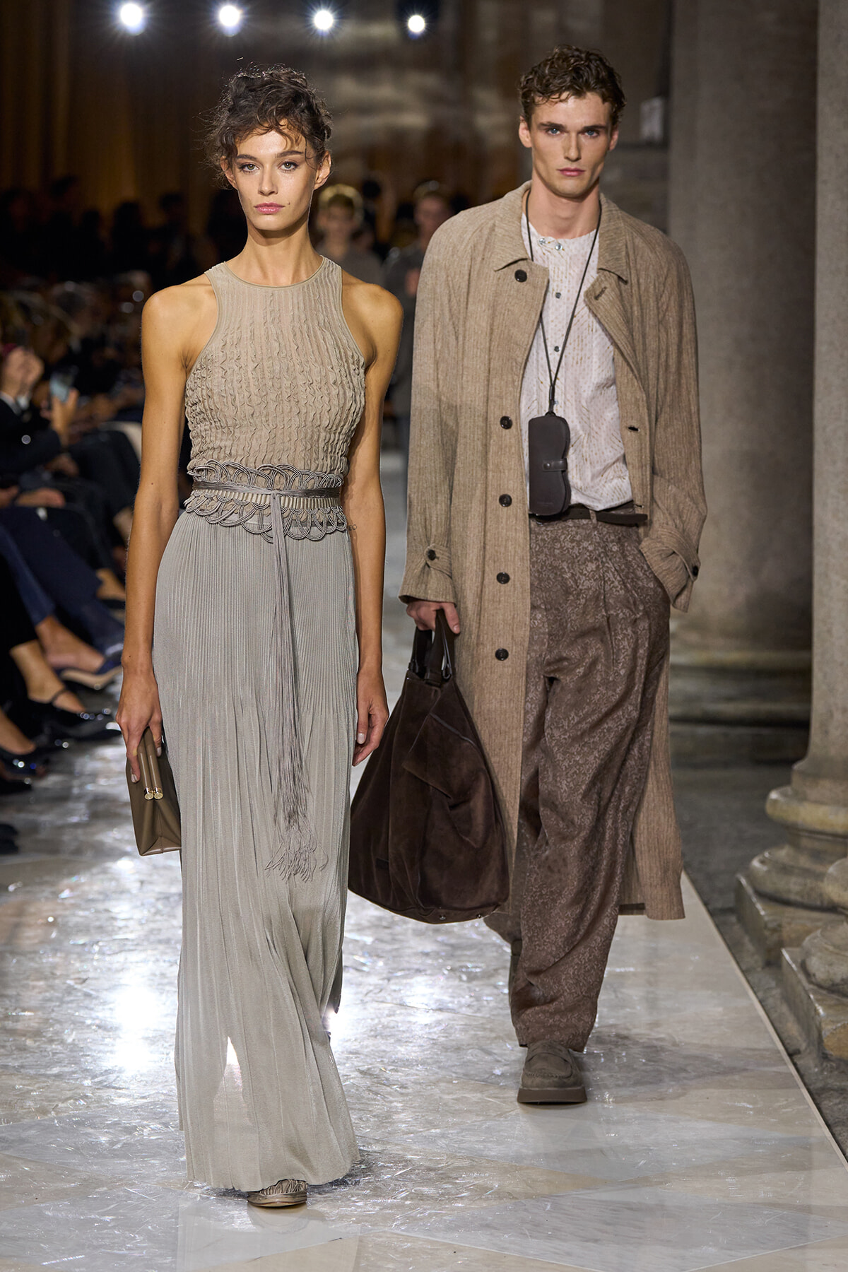 Runway models walking a fashion show: woman in a beige knit sleeveless top and a long pleated gray skirt with a decorative belt; man beside her wearing a tan overcoat, patterned brown trousers, and carrying a dark brown bag.