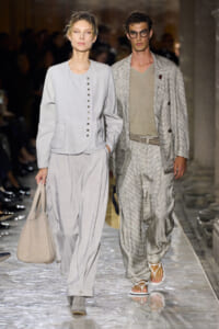 Two models walk a fashion runway in light neutral outfits; woman in pale gray buttoned jacket and wide-leg pants, man in a textured gray suit with a belt and sandals.