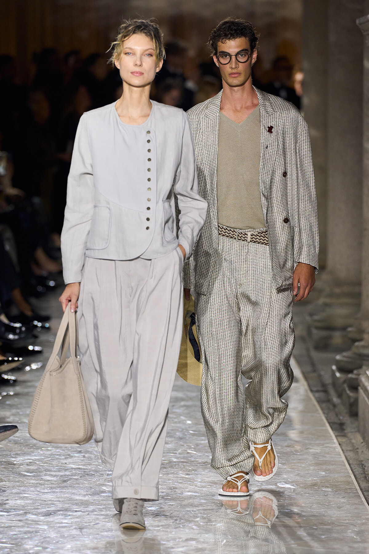 Two models walk a fashion runway in light neutral outfits; woman in pale gray buttoned jacket and wide-leg pants, man in a textured gray suit with a belt and sandals.