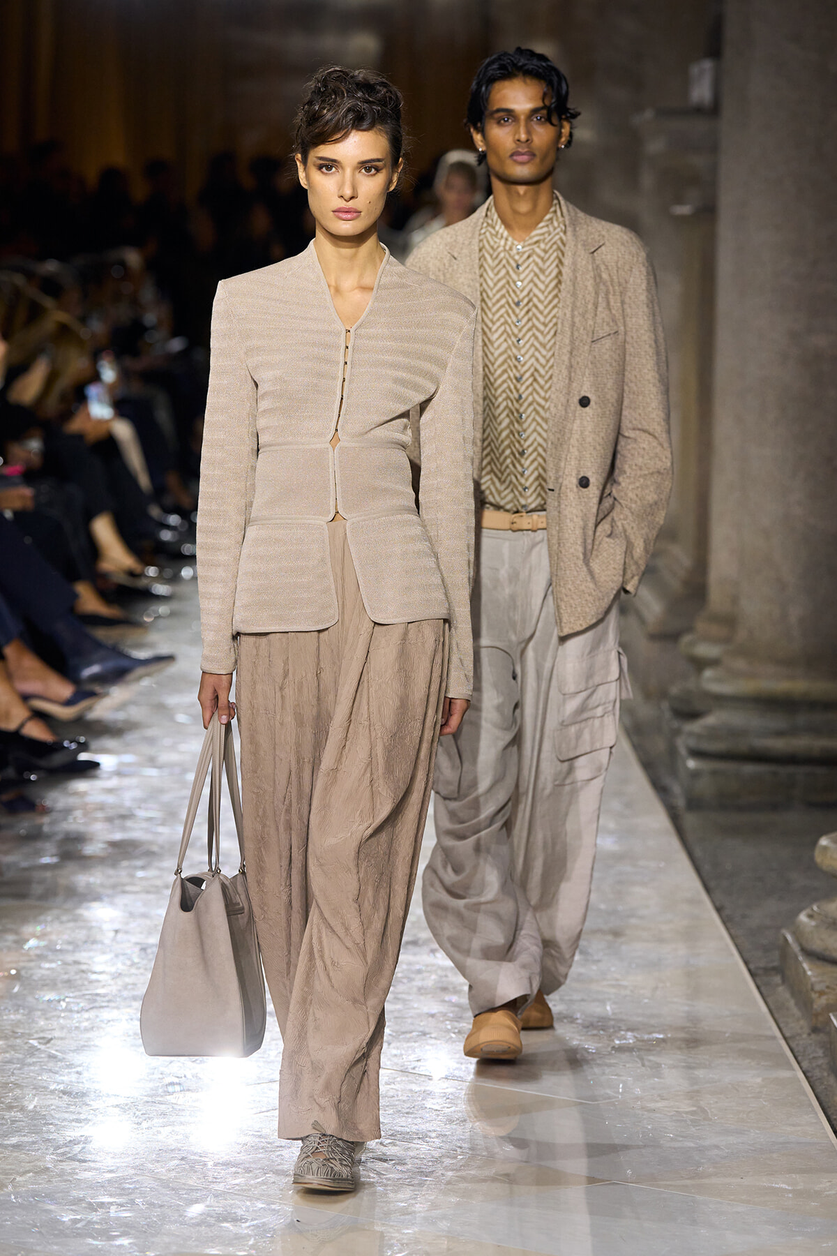 Female model walks a beige-toned outfit on a runway, carrying a light gray tote bag; she wears a structured knit jacket and wide-leg pants in soft neutrals.