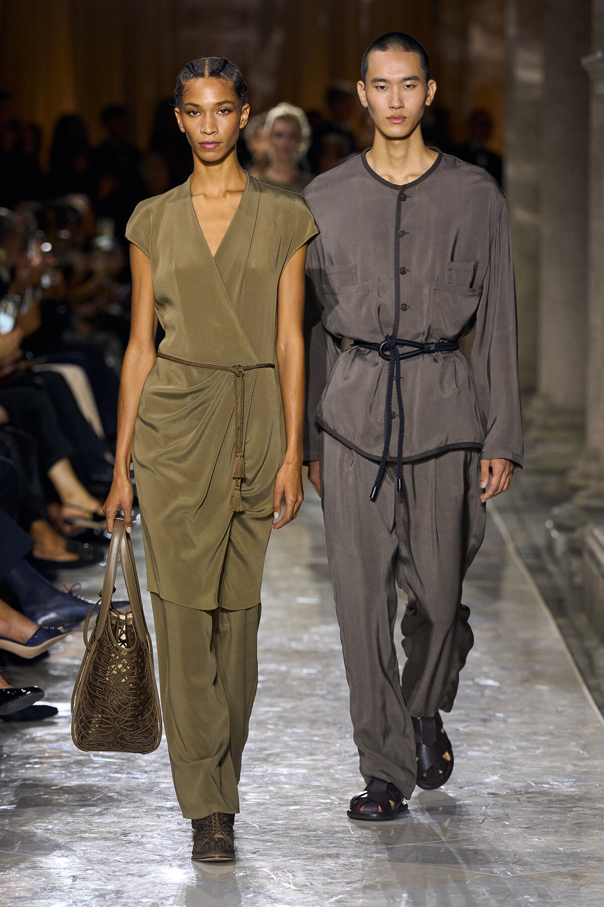 Two models walk a fashion show wearing olive and gray utility outfits with rope belts, on a glossy runway.