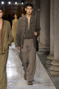 Male model walking a fashion runway in earth-toned utility jacket and wide trousers, carrying a braided bag over the shoulder near stone columns.