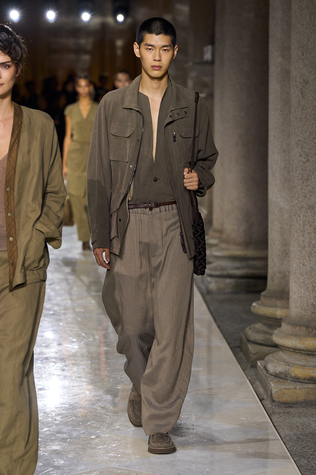 Male model walking a fashion runway in earth-toned utility jacket and wide trousers, carrying a braided bag over the shoulder near stone columns.