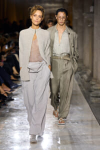 Two models walk a fashion runway in neutral-toned outfits; foreground model wears a beige cardigan over a sheer top with wide gray pants.