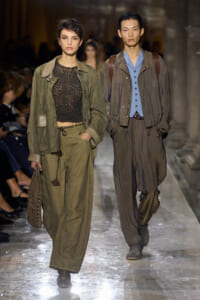 Two models walk a runway in earthy-toned outfits: woman in olive green jacket and wide pants with a black crochet top, carrying a knit bag; man in layered olive-brown jacket and blue shirt with loose trousers and shoulder bag.