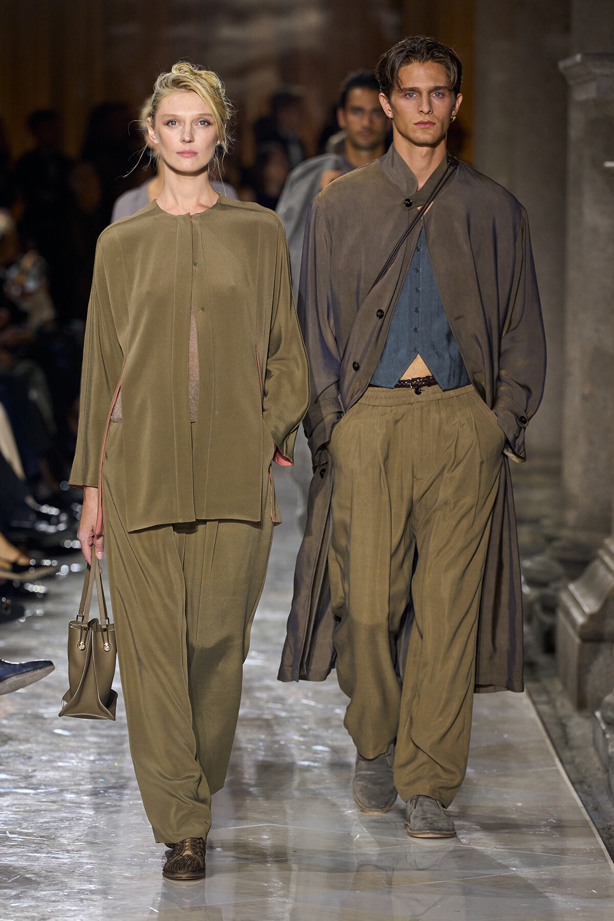 Two models walk a runway in earthy-toned outfits; the woman on the left carries a small olive handbag.