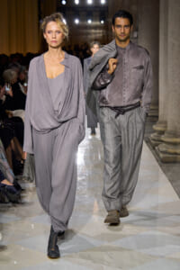 Two models walk a runway in muted gray ensembles: a woman in draped, flowing gray jumpsuit and the man in a buttoned gray shirt with matching tapered trousers, jacket slung over his shoulder.