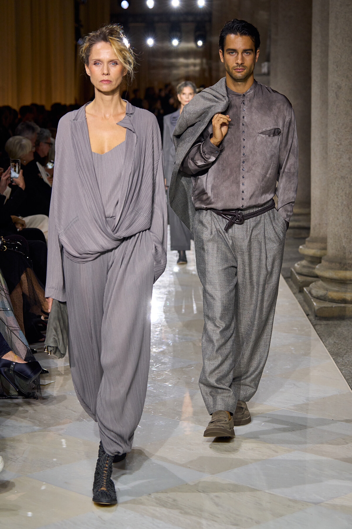Two models walk a runway in muted gray ensembles: a woman in draped, flowing gray jumpsuit and the man in a buttoned gray shirt with matching tapered trousers, jacket slung over his shoulder.