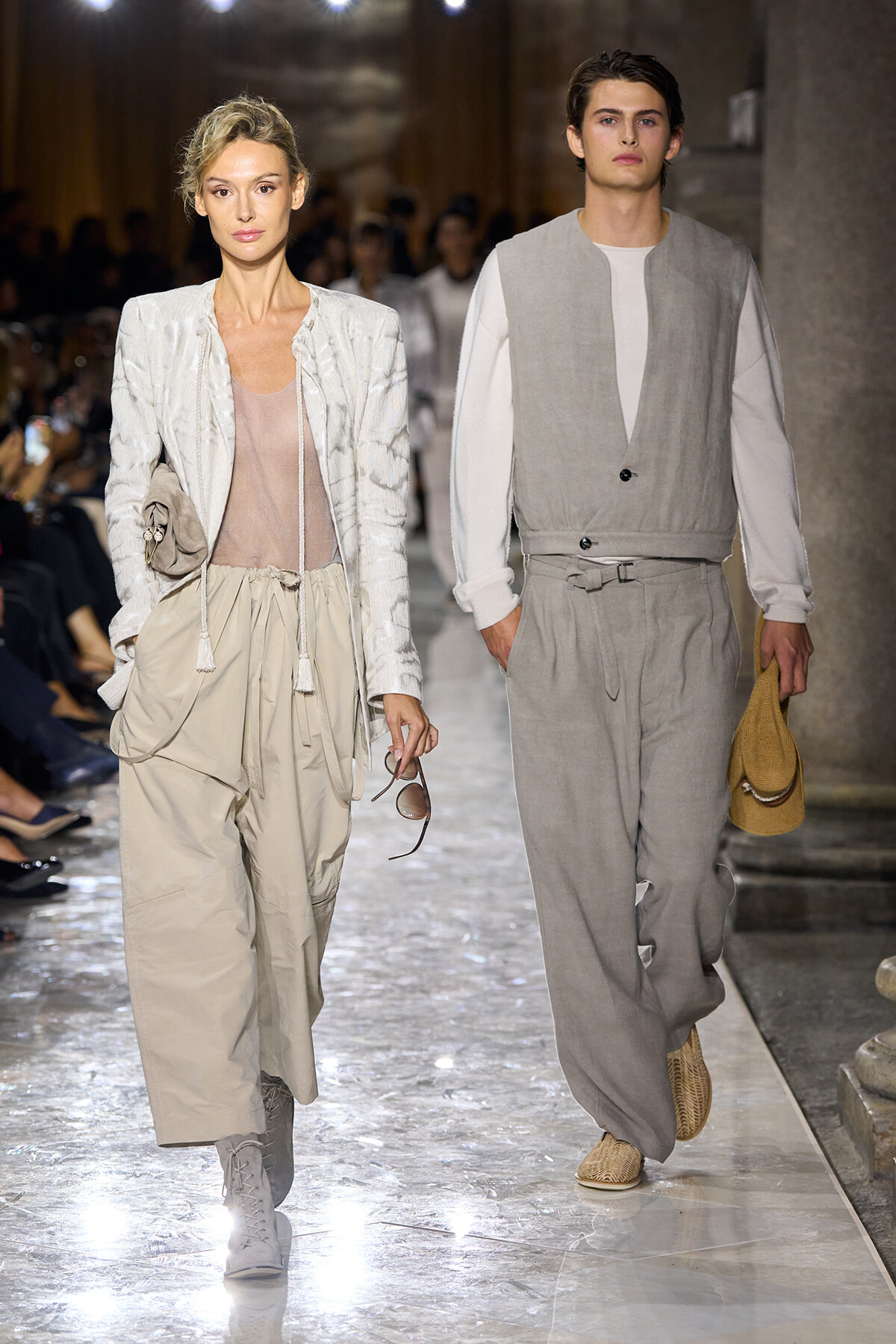 Two models walk a runway in neutral-toned outfits: a woman in a cream jacket and beige wide-leg pants with sunglasses; a man in a light gray vest and matching pants, holding a hat.
