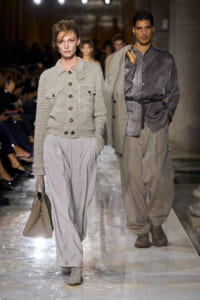 Female model walks the runway in a soft gray knit jacket with large pockets and wide gray trousers, carrying a beige handbag. A male model in a gray shirt and oversized beige coat follows behind.