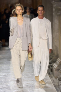 Two models walk a fashion runway in neutral beige and cream outfits, one in a textured blazer and loose pants, the other in a cream linen suit.