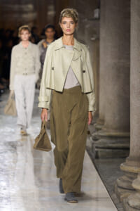Model on a runway wearing a cropped cream leather jacket, taupe blouse, olive wide-leg trousers, gray shoes, and carrying a tan handbag.