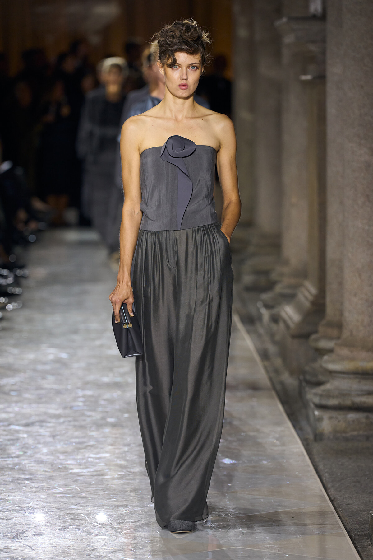 Model in a strapless charcoal gown with a fabric rose detail walks a runway, clutch in hand