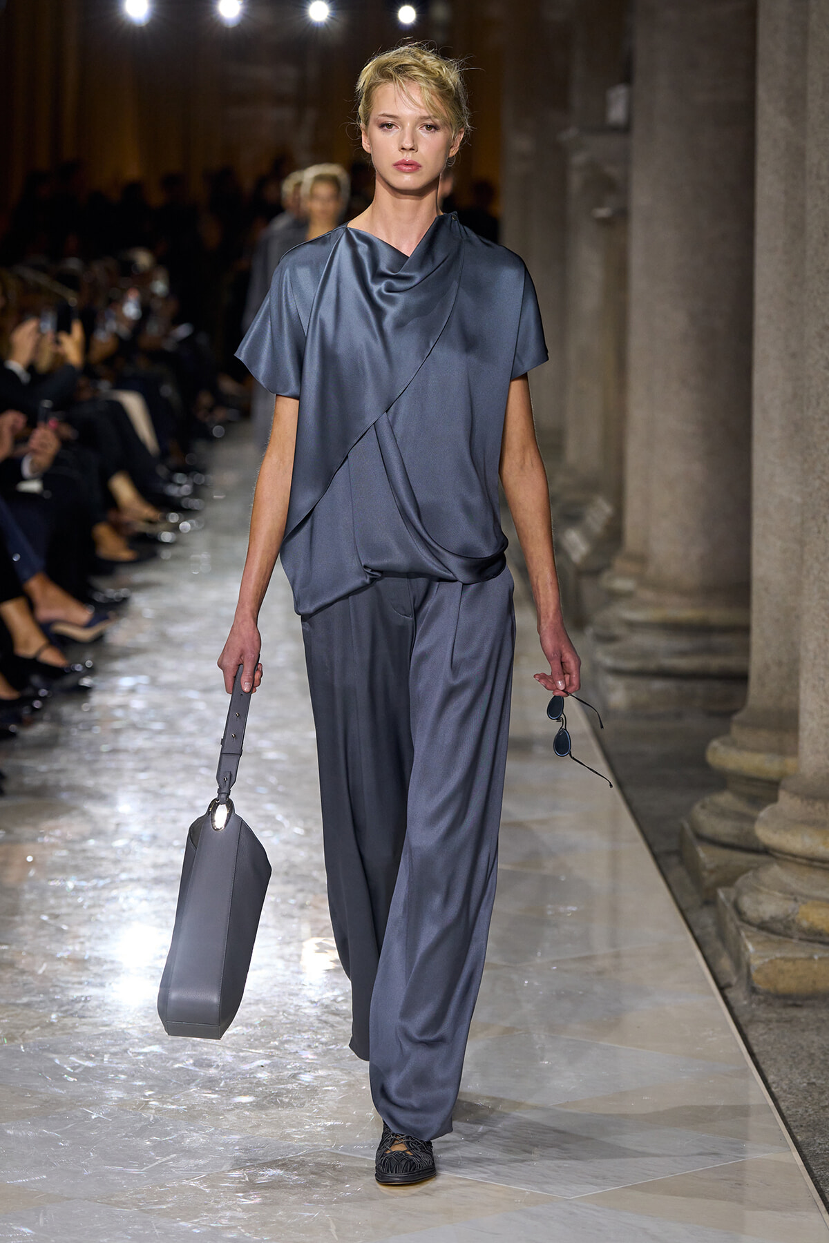 Runway model in blue satin draped top and wide-leg pants, carrying a gray rectangular bag and sunglasses.