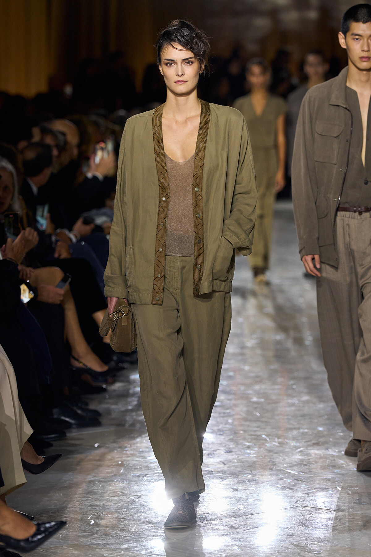 Model walking a fashion show in an olive utility jacket and matching trousers, carrying a small brown clutch.