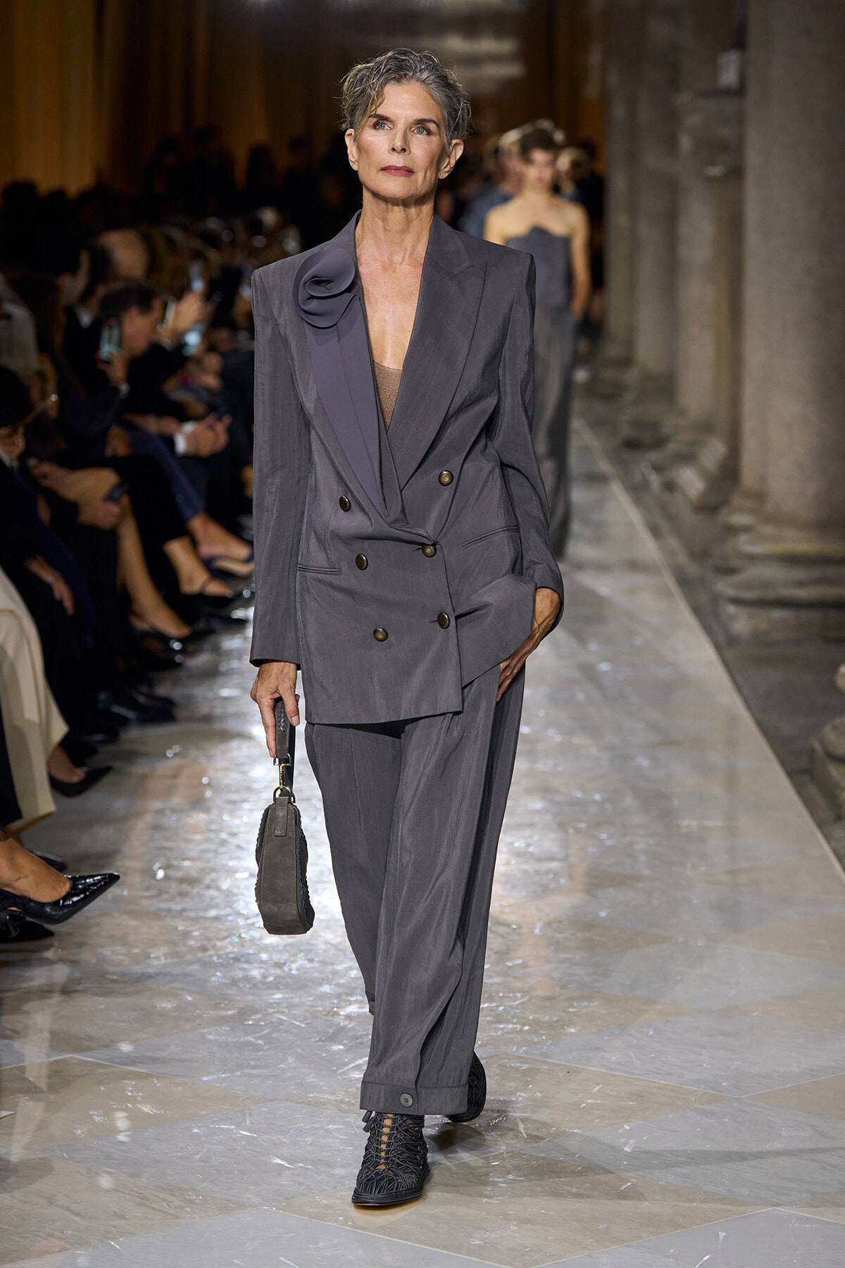 Runway model in a gray double-breasted suit with a large lapel flower, carrying a small gray handbag.