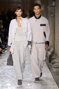 Two models walk a fashion runway in pale gray outfits; the woman carries a gray handbag and wears a light jacket with wide-leg pants.