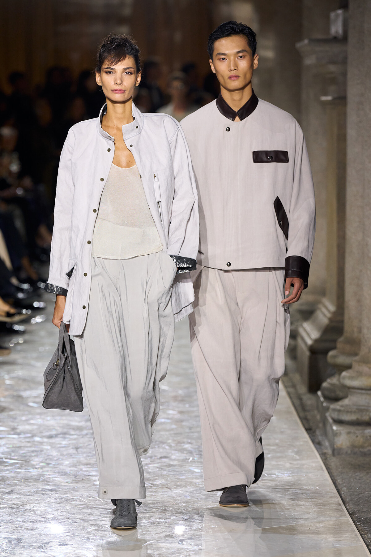 Two models walk a fashion runway in pale gray outfits; the woman carries a gray handbag and wears a light jacket with wide-leg pants.
