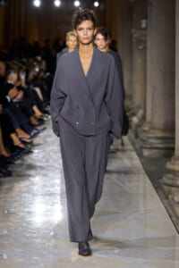 Model walks a charcoal-gray oversized blazer and matching wide-leg trousers down a fashion show runway, hands in pockets, under bright runway lights.