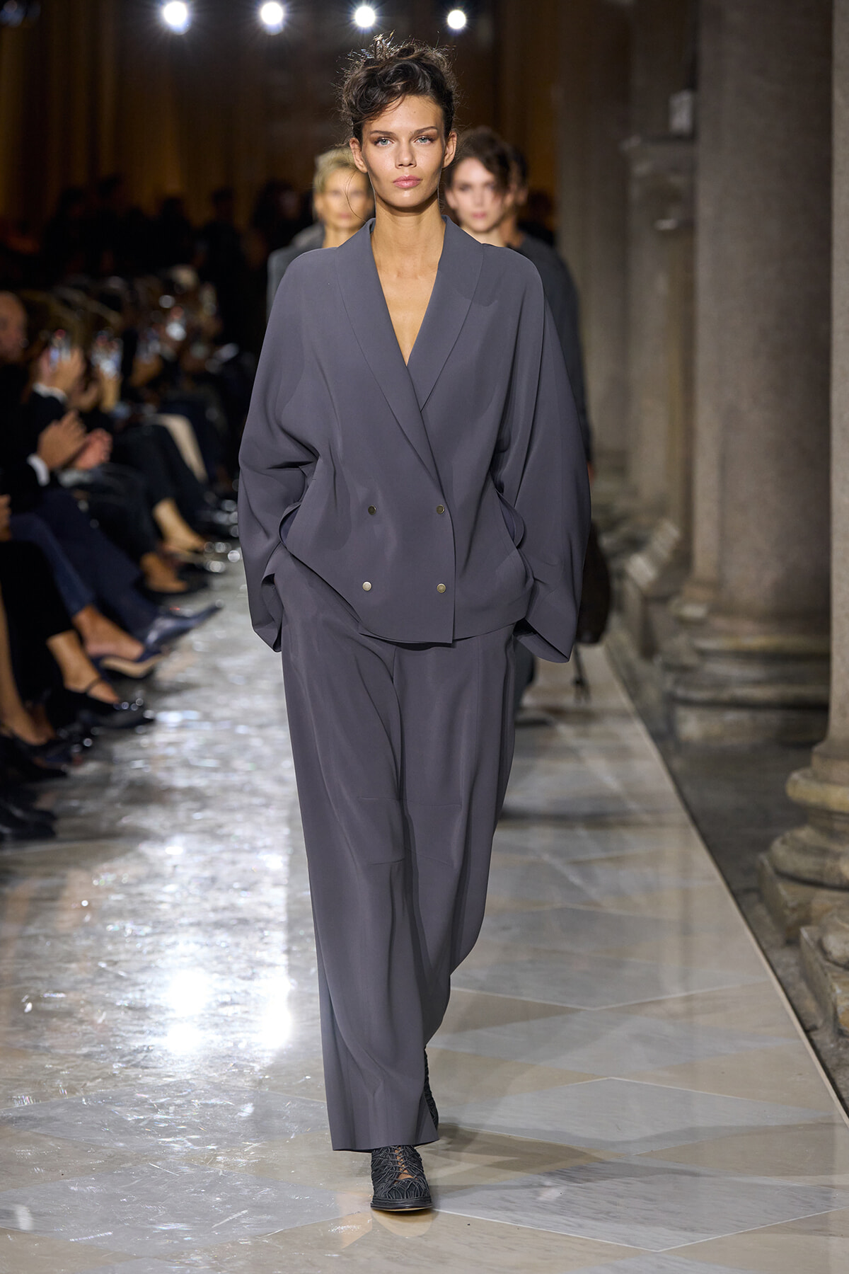 Model walks a charcoal-gray oversized blazer and matching wide-leg trousers down a fashion show runway, hands in pockets, under bright runway lights.