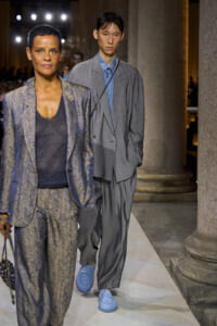 Two models walk a runway: a woman in a blue patterned suit at foreground and a man in a gray oversized suit behind her, blue shirt and blue shoes, lined columns in background.