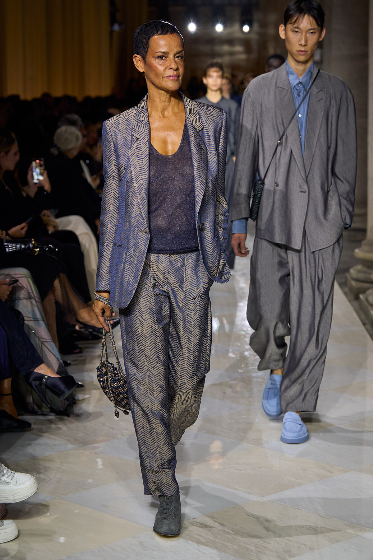 Model walks the runway in a blue chevron-pattern suit with a dark top and a small patterned handbag, audience in the background.