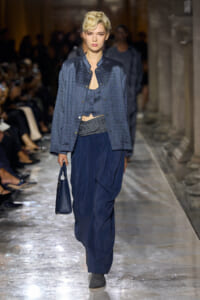 Model walks a fashion runway in a blue patterned jacket over a cropped top and draped navy pants, carrying a small blue handbag.