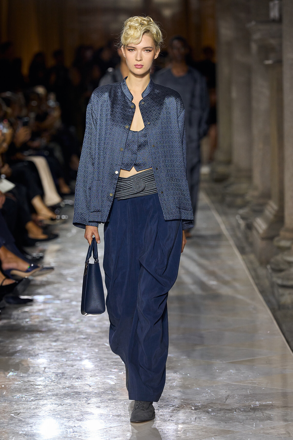 Model walks a fashion runway in a blue patterned jacket over a cropped top and draped navy pants, carrying a small blue handbag.