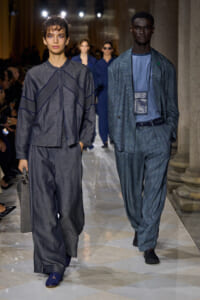 Two models walk a runway in blue-gray outfits; left is in a loose gray top and wide pants carrying a gray bag, right in a patterned blue suit with a matching shirt.