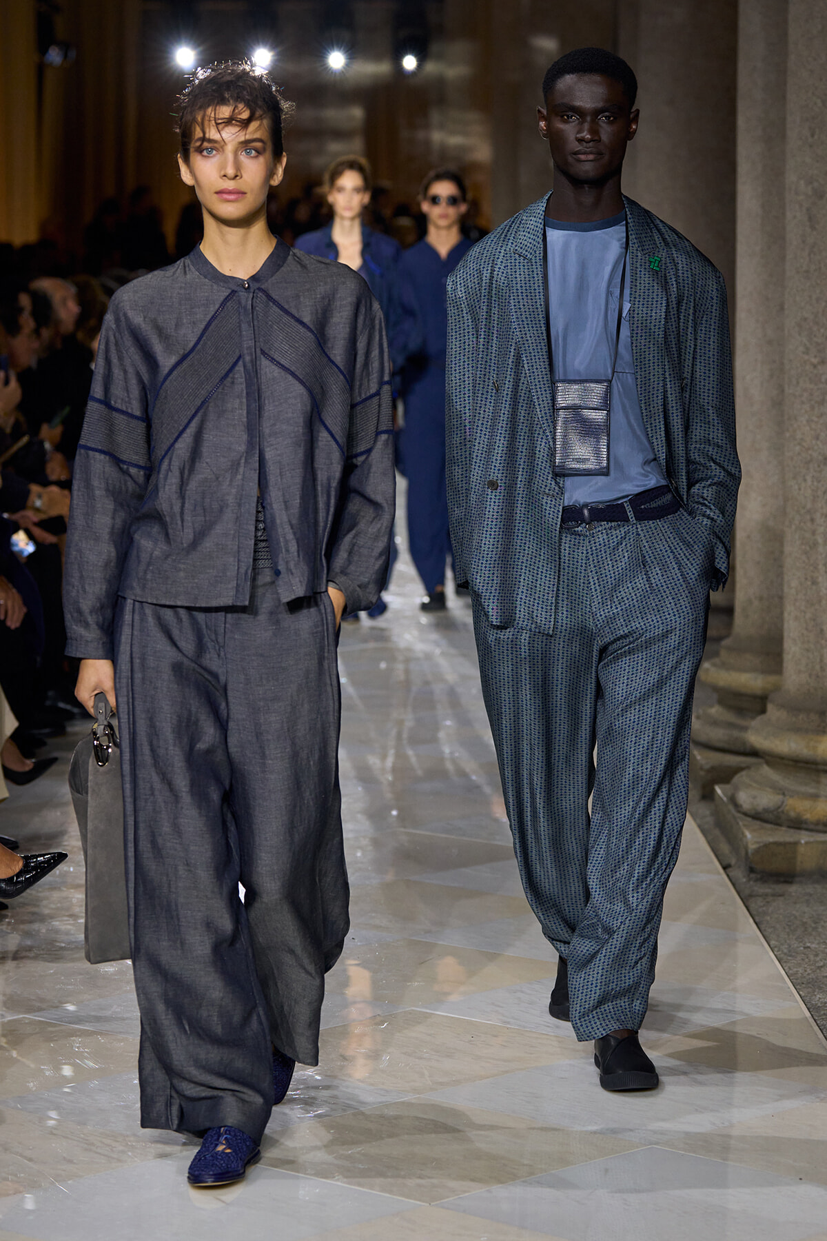 Two models walk a runway in blue-gray outfits; left is in a loose gray top and wide pants carrying a gray bag, right in a patterned blue suit with a matching shirt.