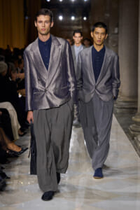 Two male models walk a runway in shimmering gray blazers, dark shirts, and oversized gray trousers, with an audience visible on the left.