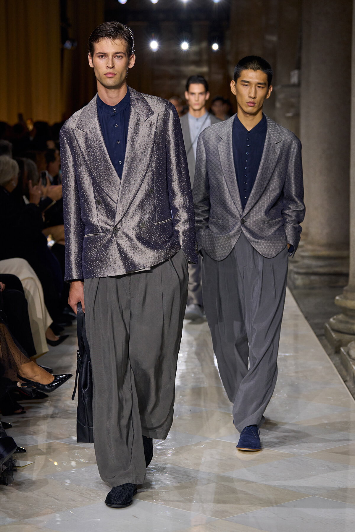 Two male models walk a runway in shimmering gray blazers, dark shirts, and oversized gray trousers, with an audience visible on the left.