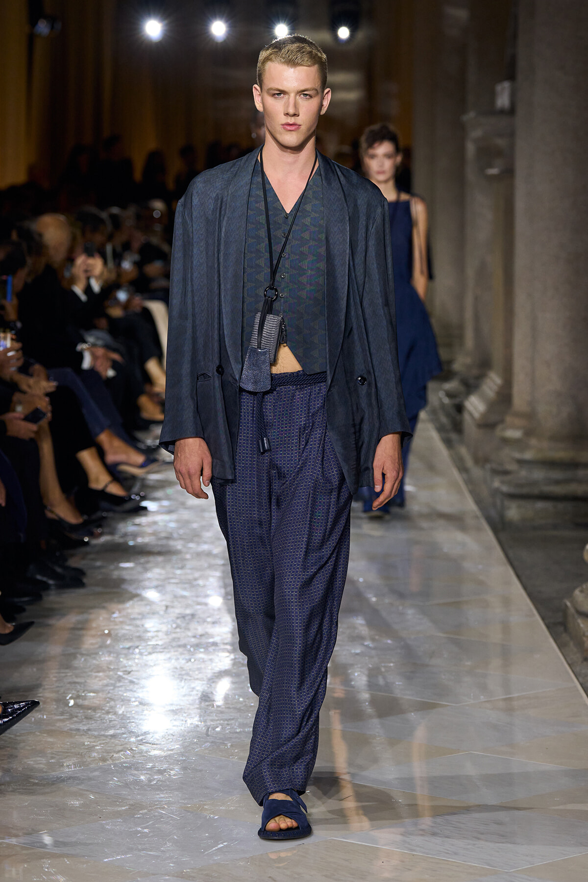 Male model on a fashion runway wearing a navy blue layered outfit: loose blazer, patterned vest, oversized pants, and blue sandals; audience visible in the background.